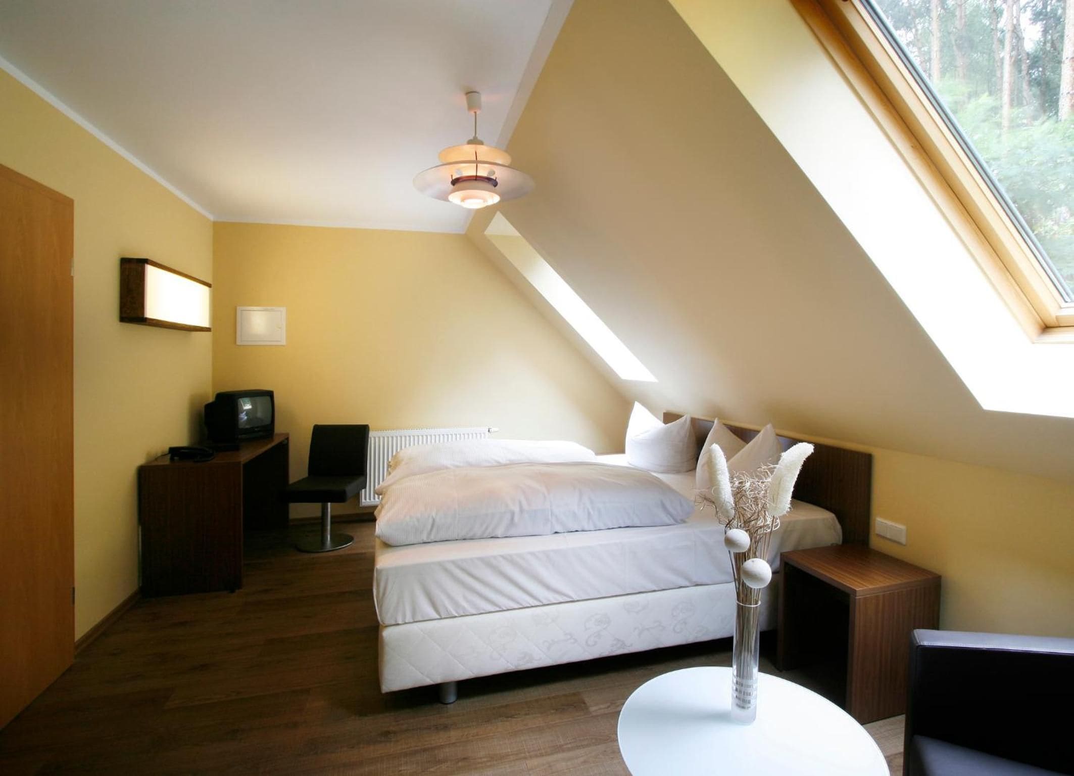 A bedroom with a bed, bedside tables, a desk, a chair, a television, and a window with a view of trees.