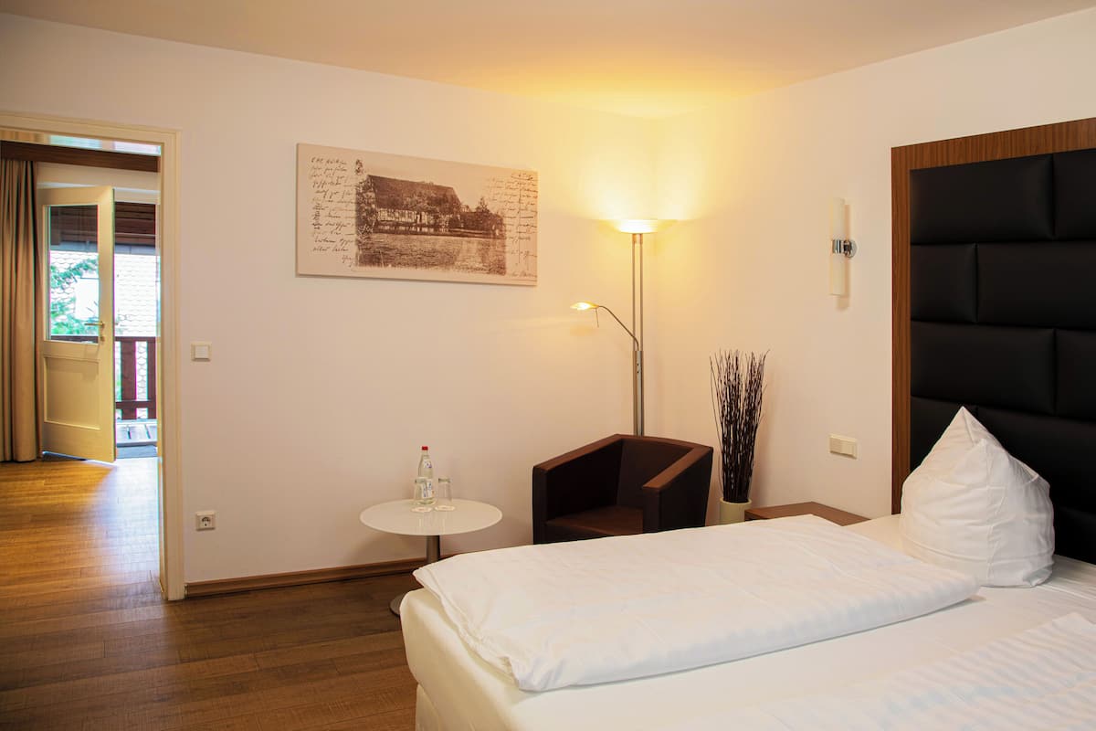Habitación de hotel con cama, una mesita con una botella y vasos, una silla, una lámpara de pared y un cuadro.