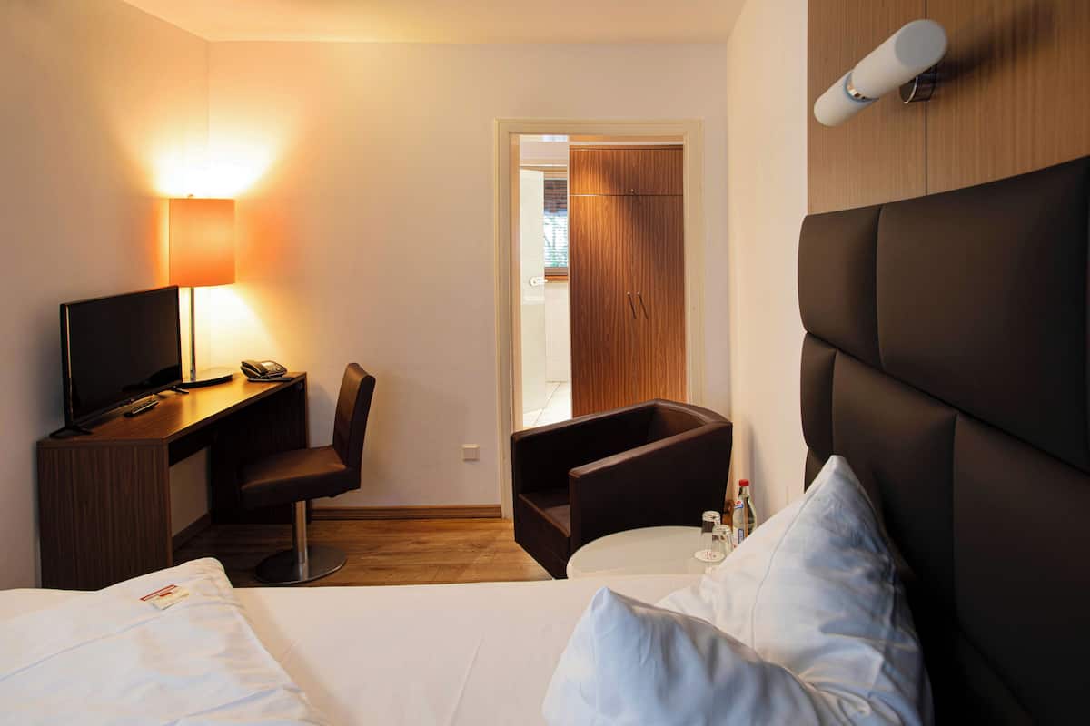Una habitación de hotel con una cama, un escritorio con una silla, un televisor, una lámpara y una mesita de noche con una botella.