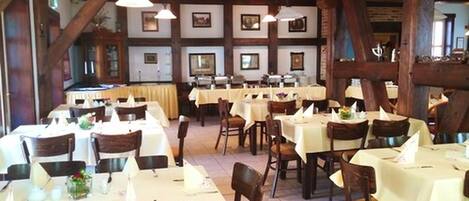 A restaurant with multiple tables and chairs, wooden beams on the ceiling, and framed pictures on the walls.