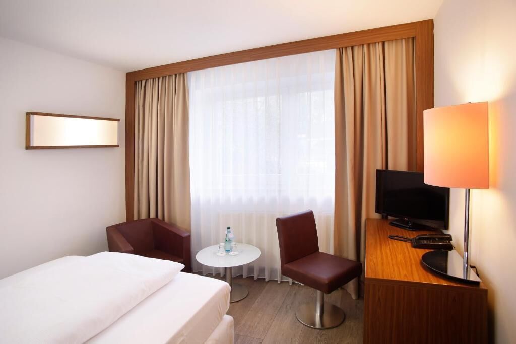 Ein Hotelzimmer mit einem Bett, zwei Stühlen, einem kleinen Tisch mit einer Flasche, einem Fernseher und einer Lampe.