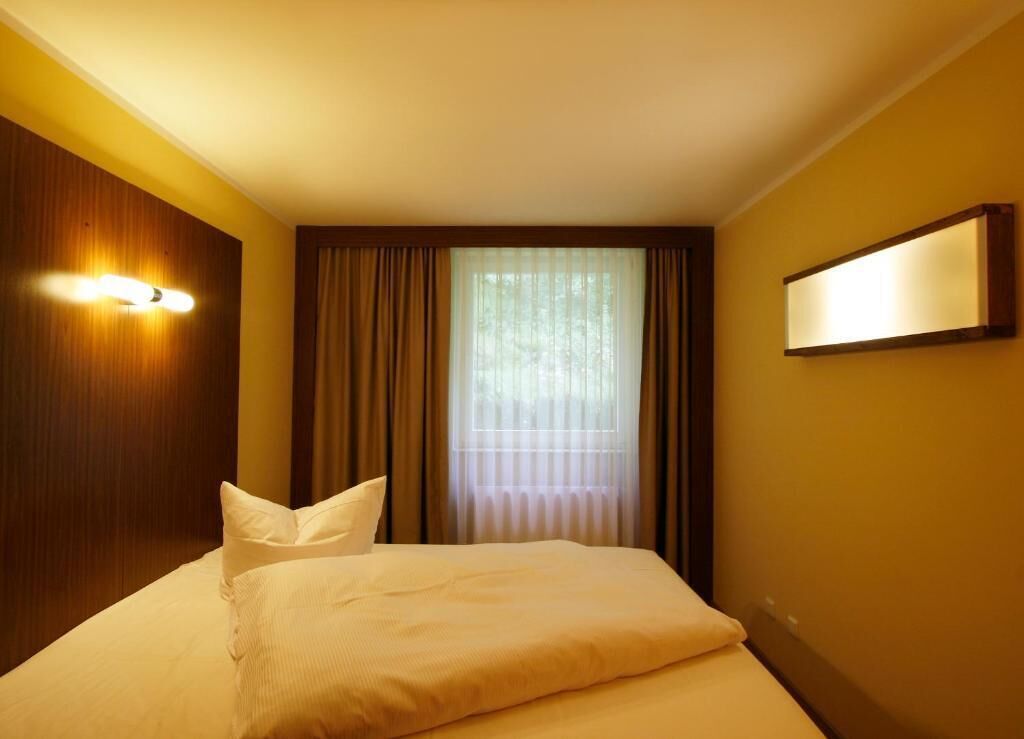 Een hotelkamer met een bed, gordijnen, een raam met zonwering en een wandlamp.