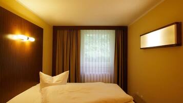 Een hotelkamer met een bed, gordijnen, een raam met zonwering en een wandlamp.