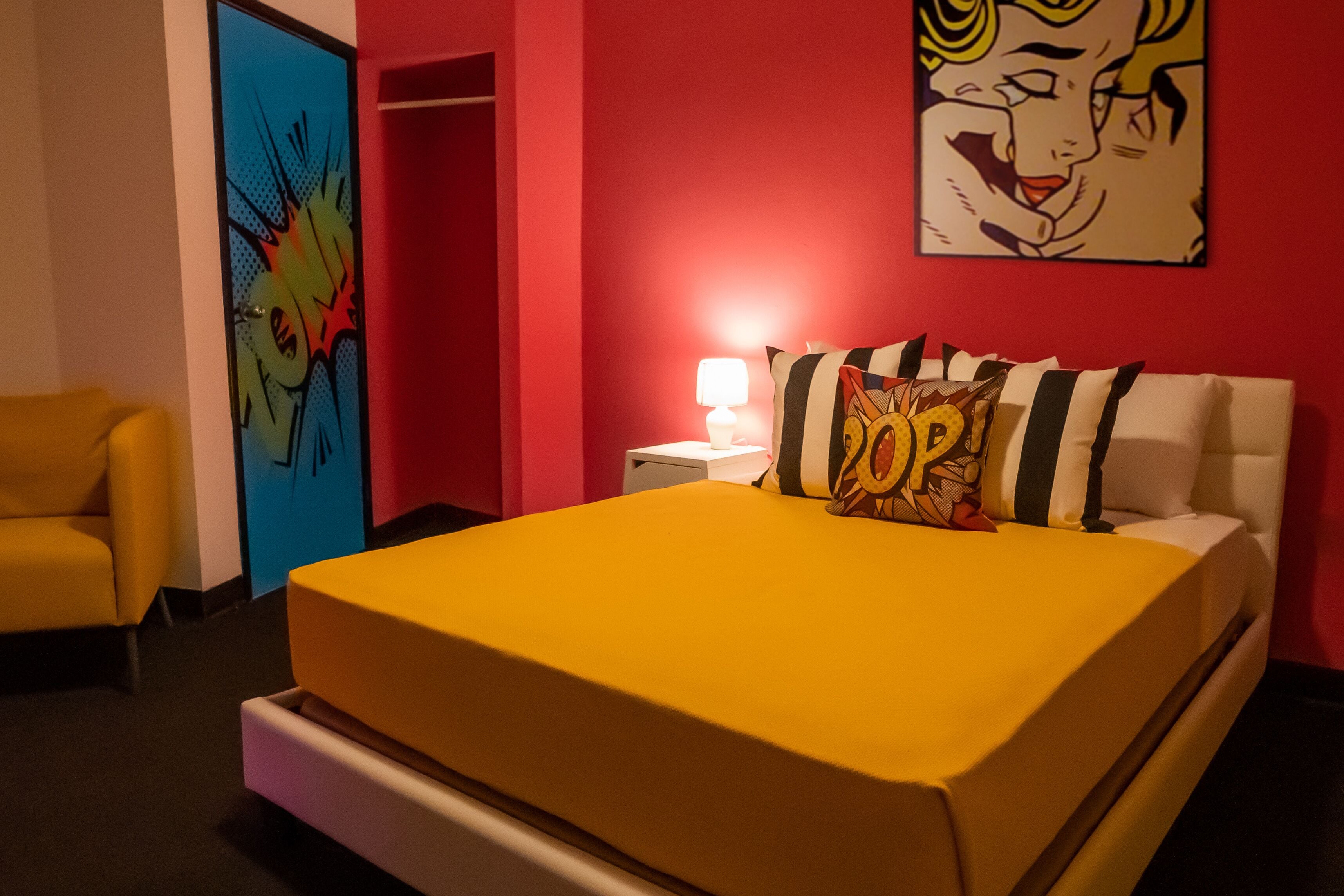 A hotel room with a yellow bed, a red wall, a blue door, and a yellow armchair.