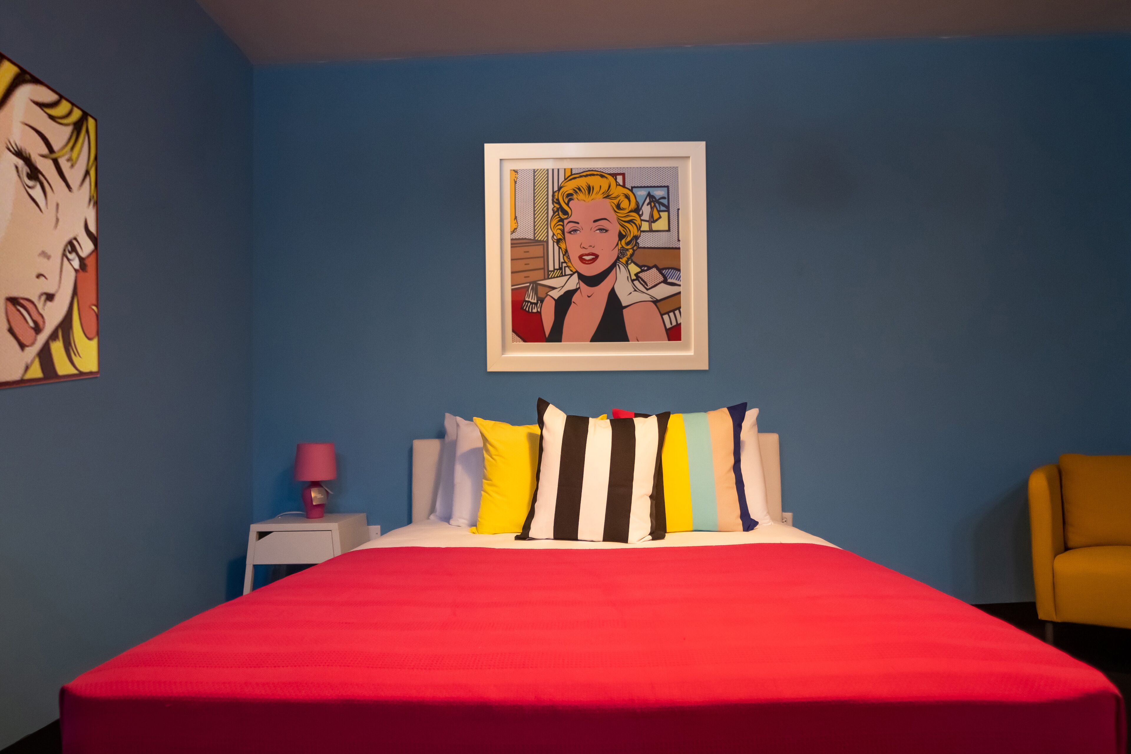 A room with a red bedspread, a framed pop art painting, and a yellow armchair.