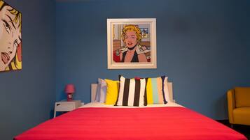 A room with a red bedspread, a framed pop art painting, and a yellow armchair.