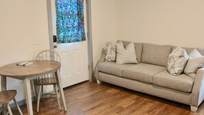 A living room with a beige sofa, patterned cushions, a wooden dining table with two chairs, and a window with a floral curtain. - Arctic Dragonfly Inn (Healy)