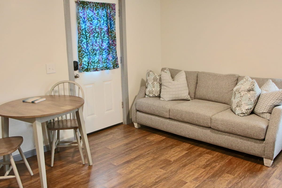 A living room with a beige sofa, patterned cushions, a wooden dining table with two chairs, and a window with a floral curtain.