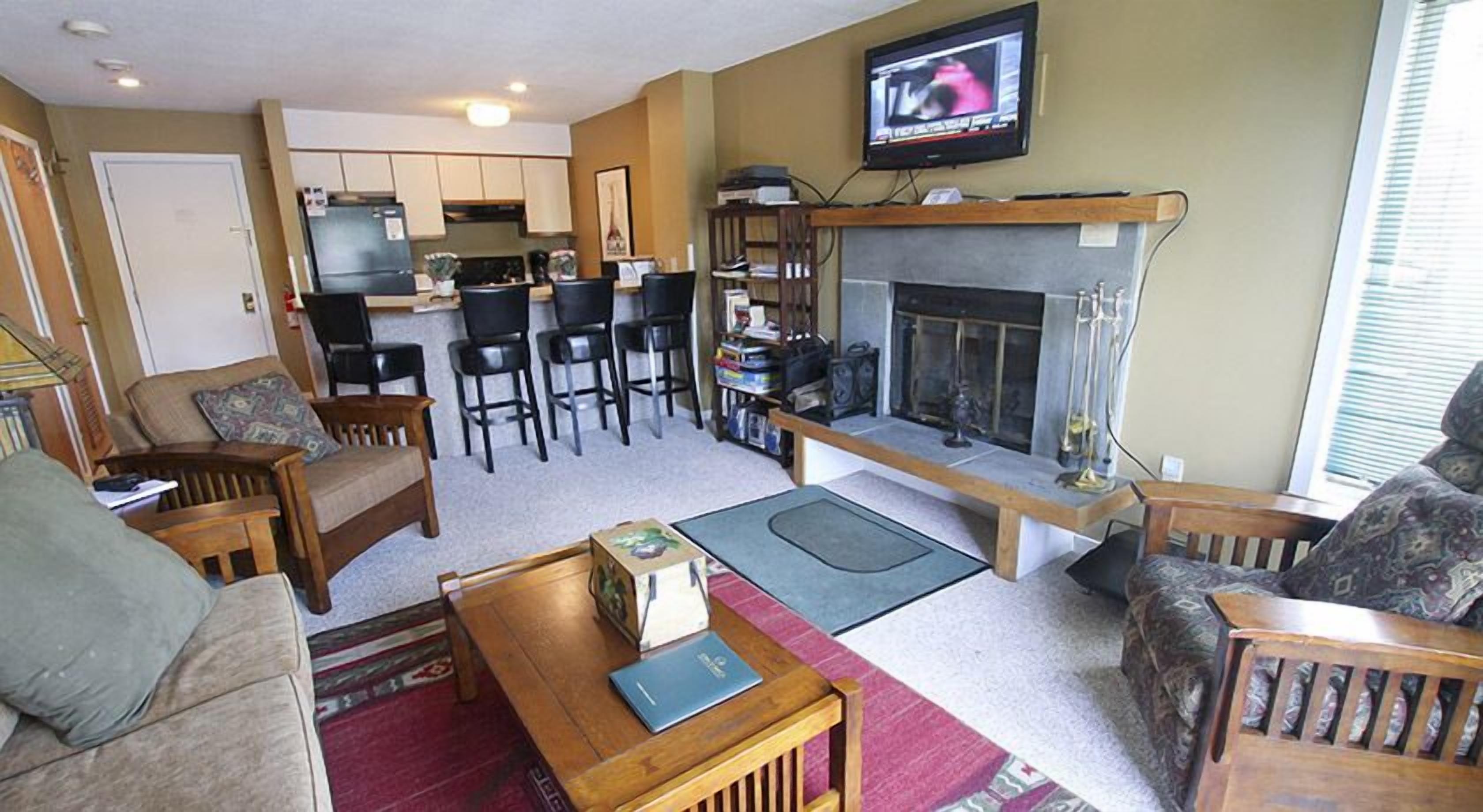 A living room with a fireplace, a television, and a kitchen area with chairs and a refrigerator.