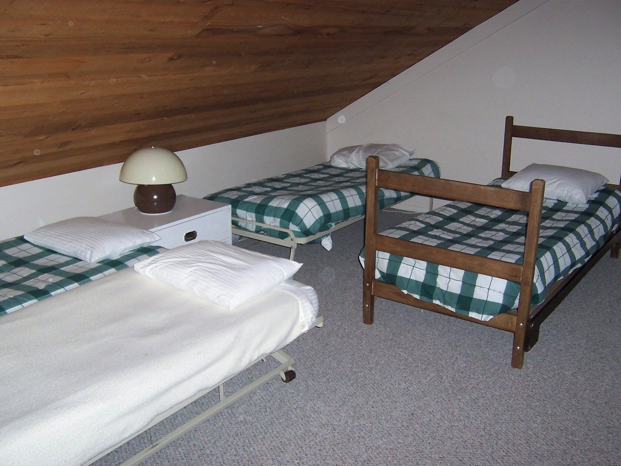 Una habitación pequeña y con poca luz, con tres literas, una mesita de noche individual con una lámpara y un techo de madera.