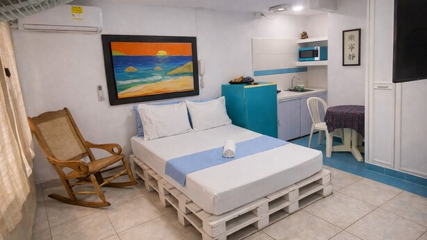 A room with a bed on pallets, a rocking chair, a desk, a chair, a microwave, and a painting of a sunset.