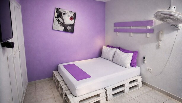 A bedroom with a bed on pallets, a purple wall, and a painting. - Quillahost Guesthouse (Barranquilla)