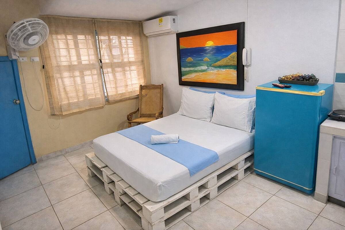 A room with a bed on pallets, a chair, a fan, a painting, and a phone.