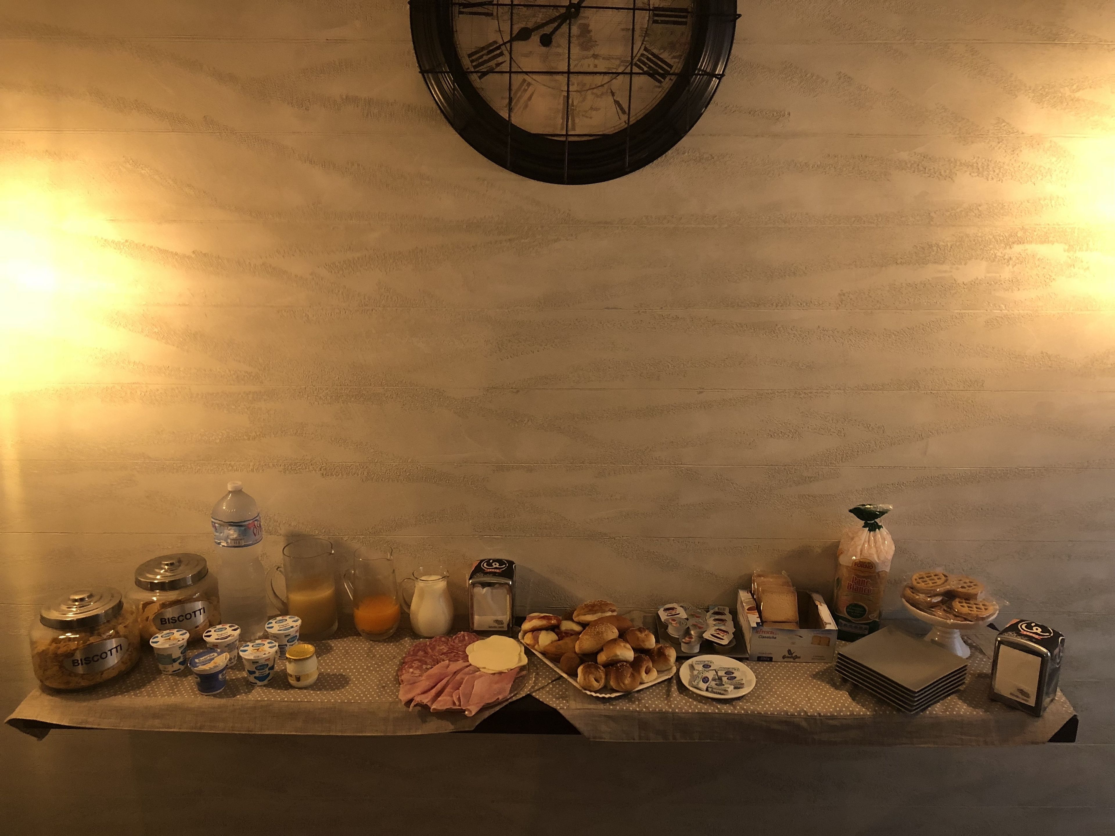 A table with various food items and condiments, including jars labeled 'BISCUCCI', a plate of pastries, and a clock on the wall.