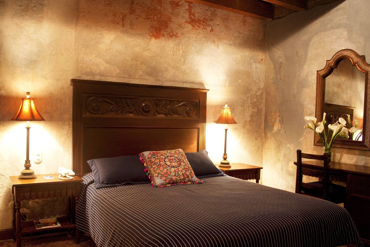 A bedroom with a large bed, two bedside tables, a nightstand, a mirror, and a vase with flowers.