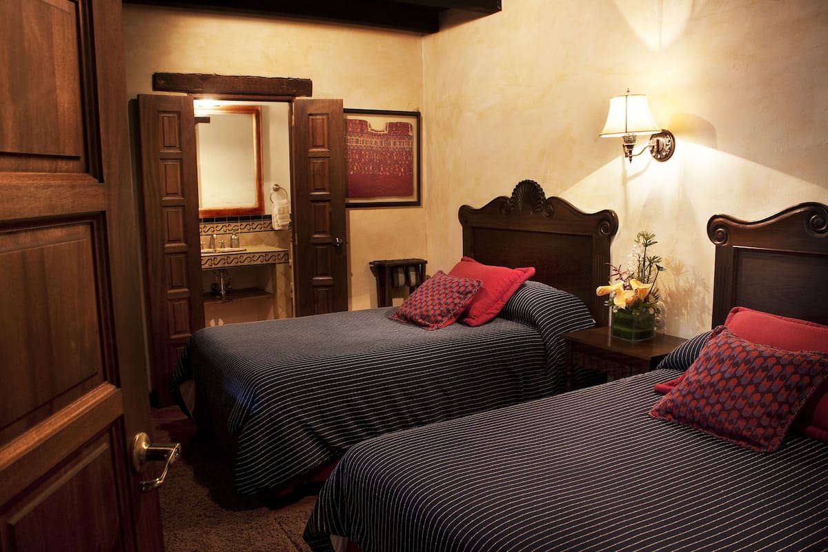 A hotel room with two beds, a wooden door, a mirror, a nightstand with a lamp, and a wall-mounted picture.