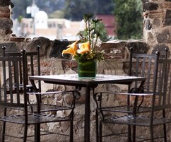 Una mesa con un jarrón de flores, dos sillas y un muro de piedra al fondo.