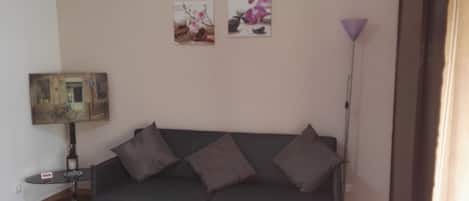 A living room with a dark green sofa, four cushions, a small round table with a lamp, and two wall-mounted pictures.