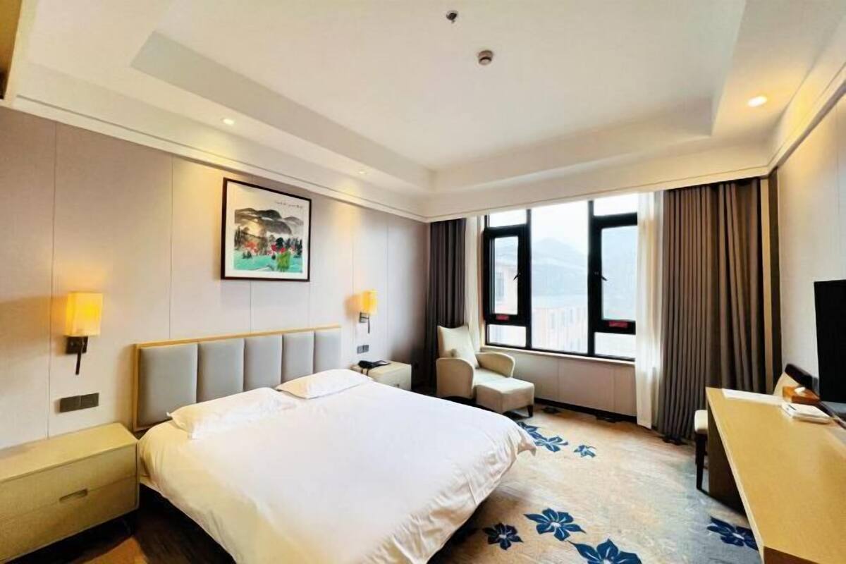 Une chambre d’hôtel comprenant un lit, des tables de chevet, un bureau, une chaise, une fenêtre avec des rideaux et un tableau au mur.