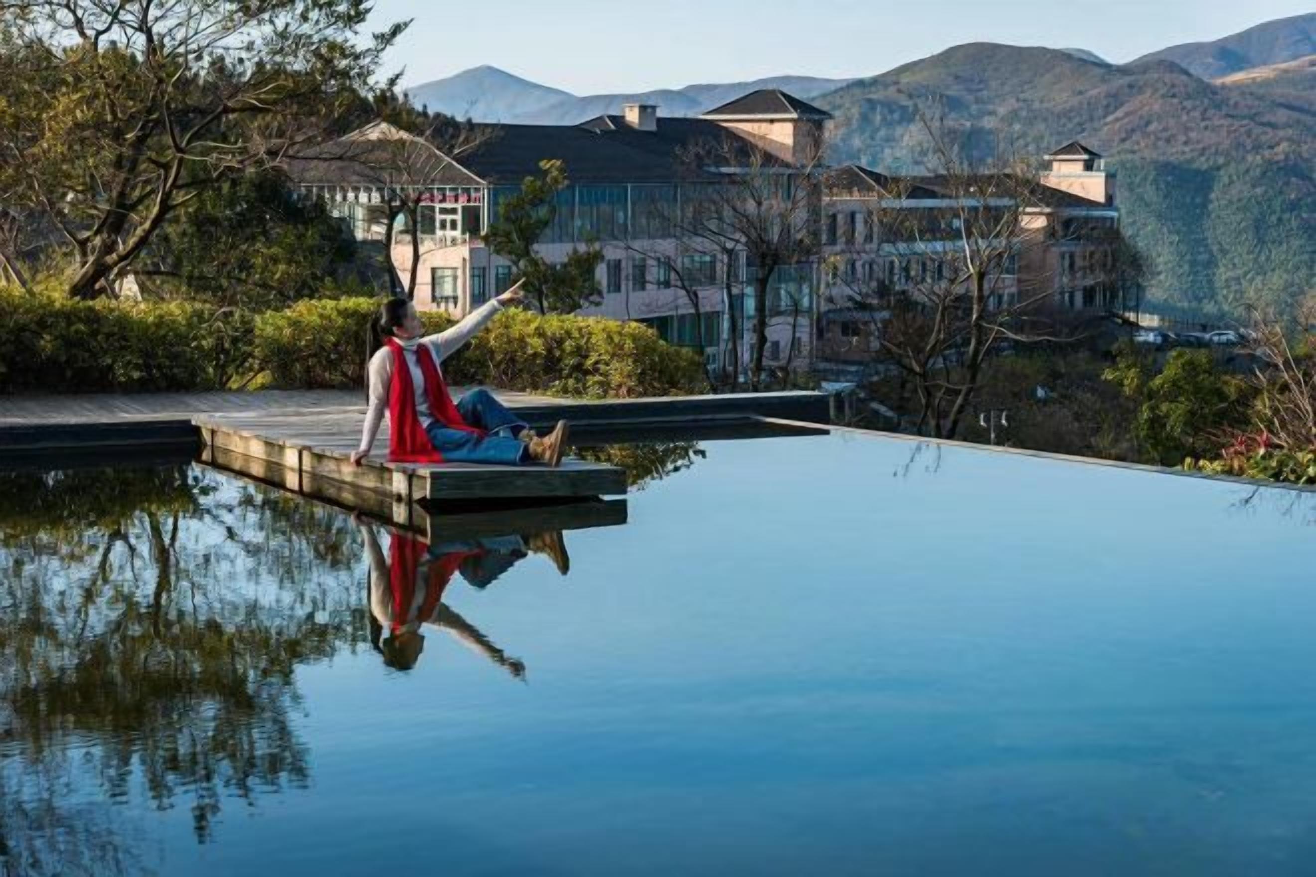 Una persona sentada en un muelle que se extiende sobre una masa de agua tranquila, con edificios y montañas al fondo.