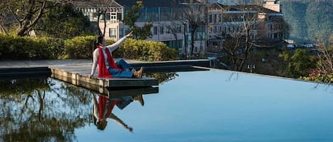 Une personne est assise sur une jetée qui s’avance sur une étendue d’eau calme, avec des bâtiments et des montagnes en arrière-plan.