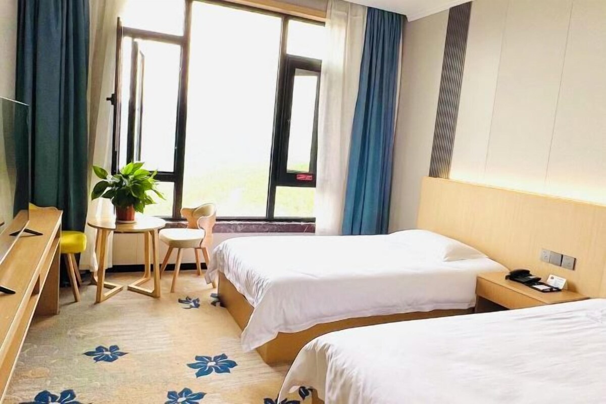 A hotel room with two beds, a desk with a chair, a small table with a plant, and a window with curtains.