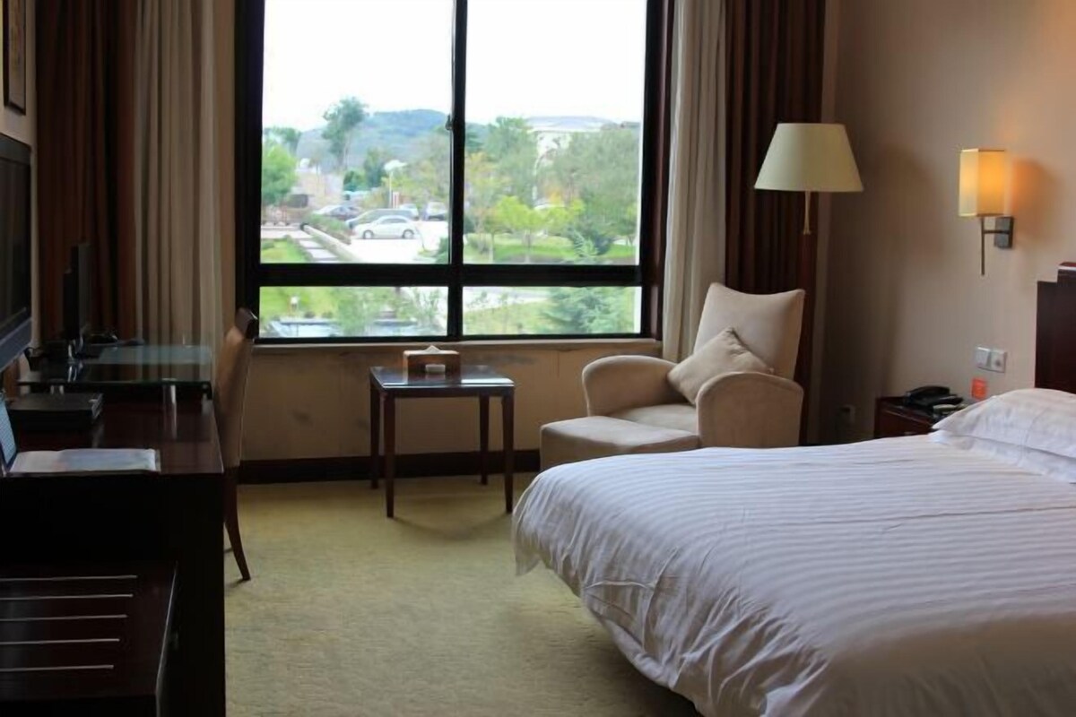 Une chambre d’hôtel comprenant un lit, un bureau, une chaise, une petite table et une fenêtre donnant sur un parking et des espaces verts.