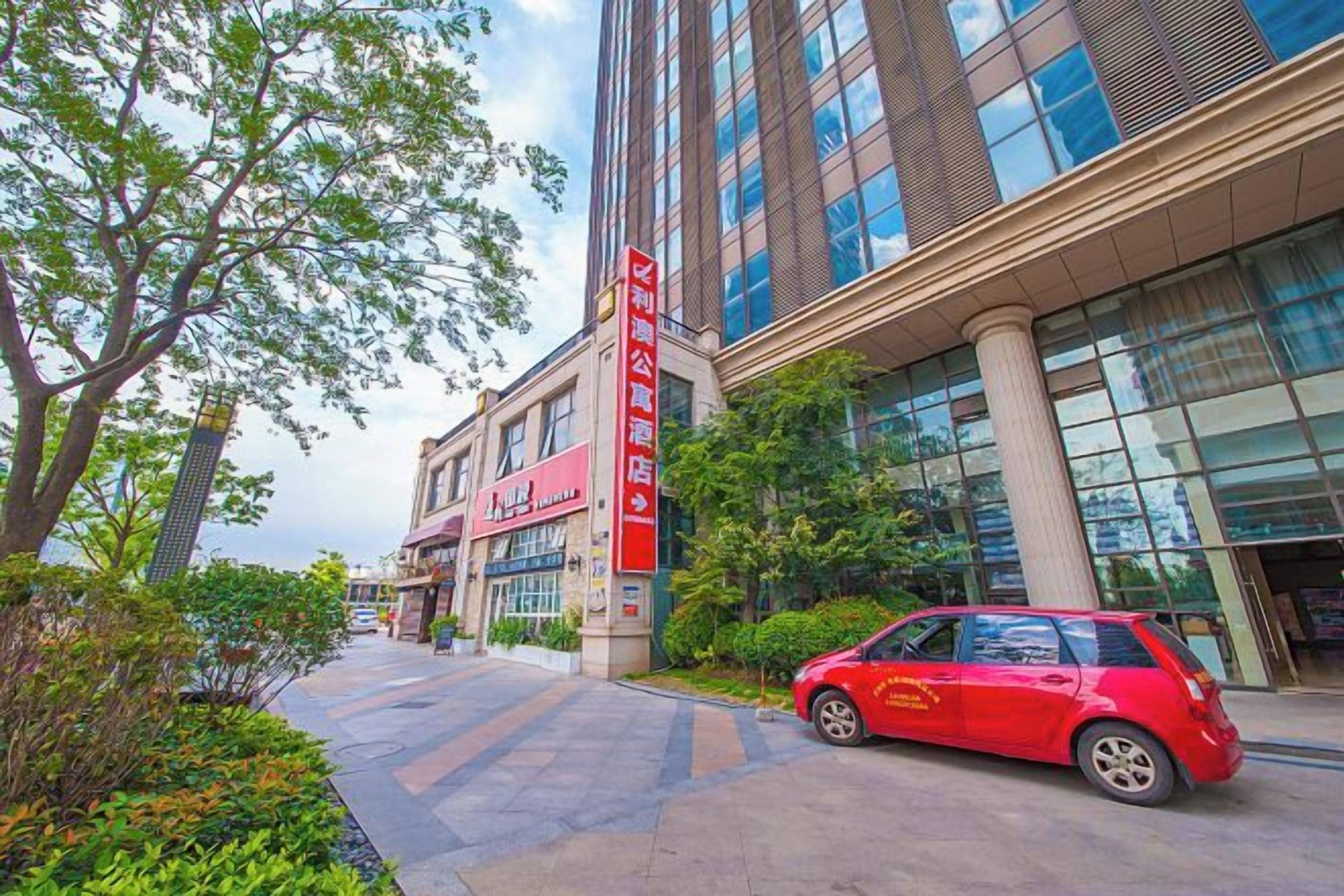 Un edificio moderno con un auto rojo estacionado adelante, cielo despejado y un camino bordeado de árboles.