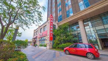 Un edificio moderno con un auto rojo estacionado adelante, cielo despejado y un camino bordeado de árboles.