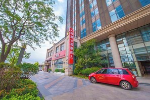 Un edificio moderno con un auto rojo estacionado adelante, cielo despejado y un camino bordeado de árboles.