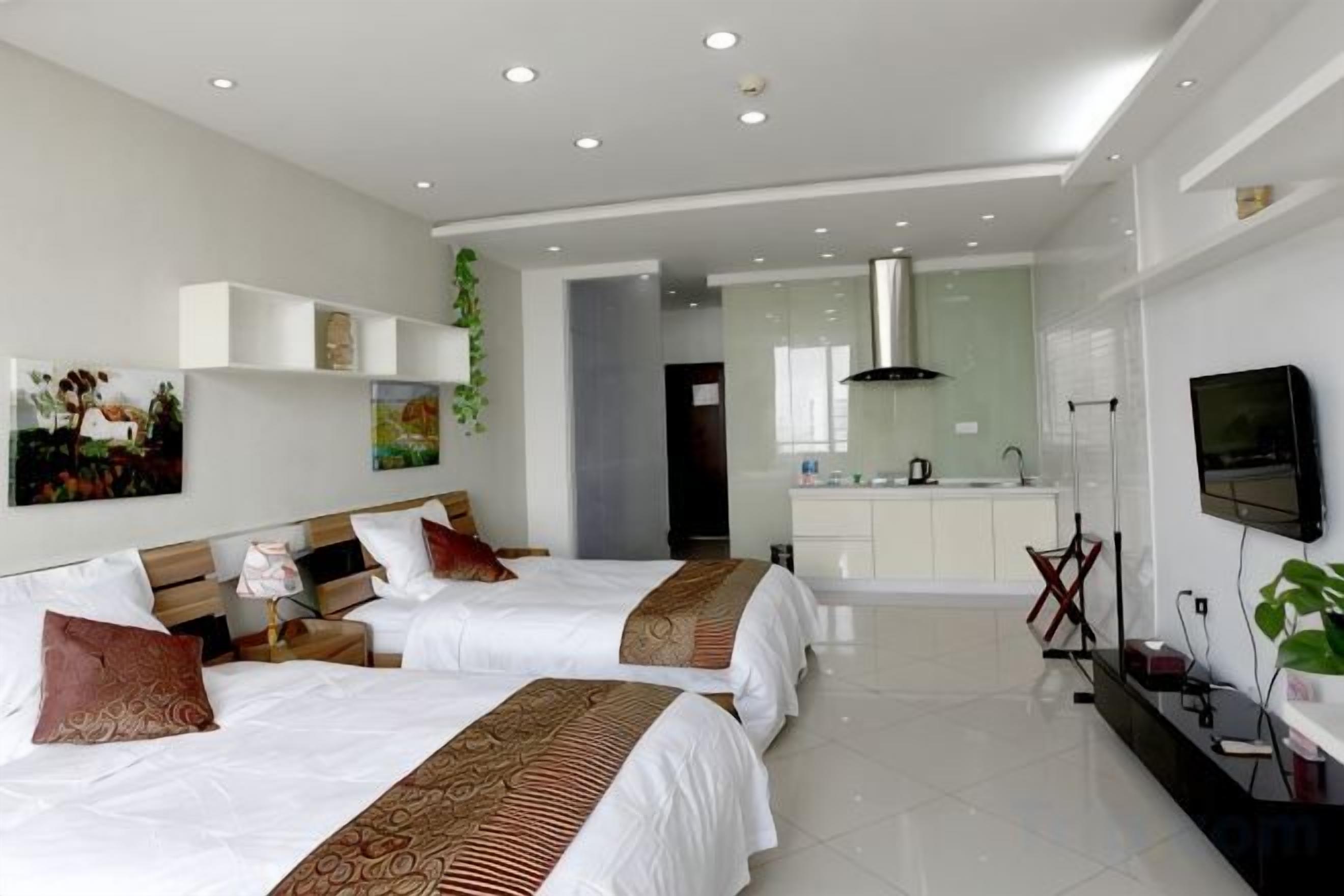A modern hotel room with two beds, a kitchenette, and a TV.