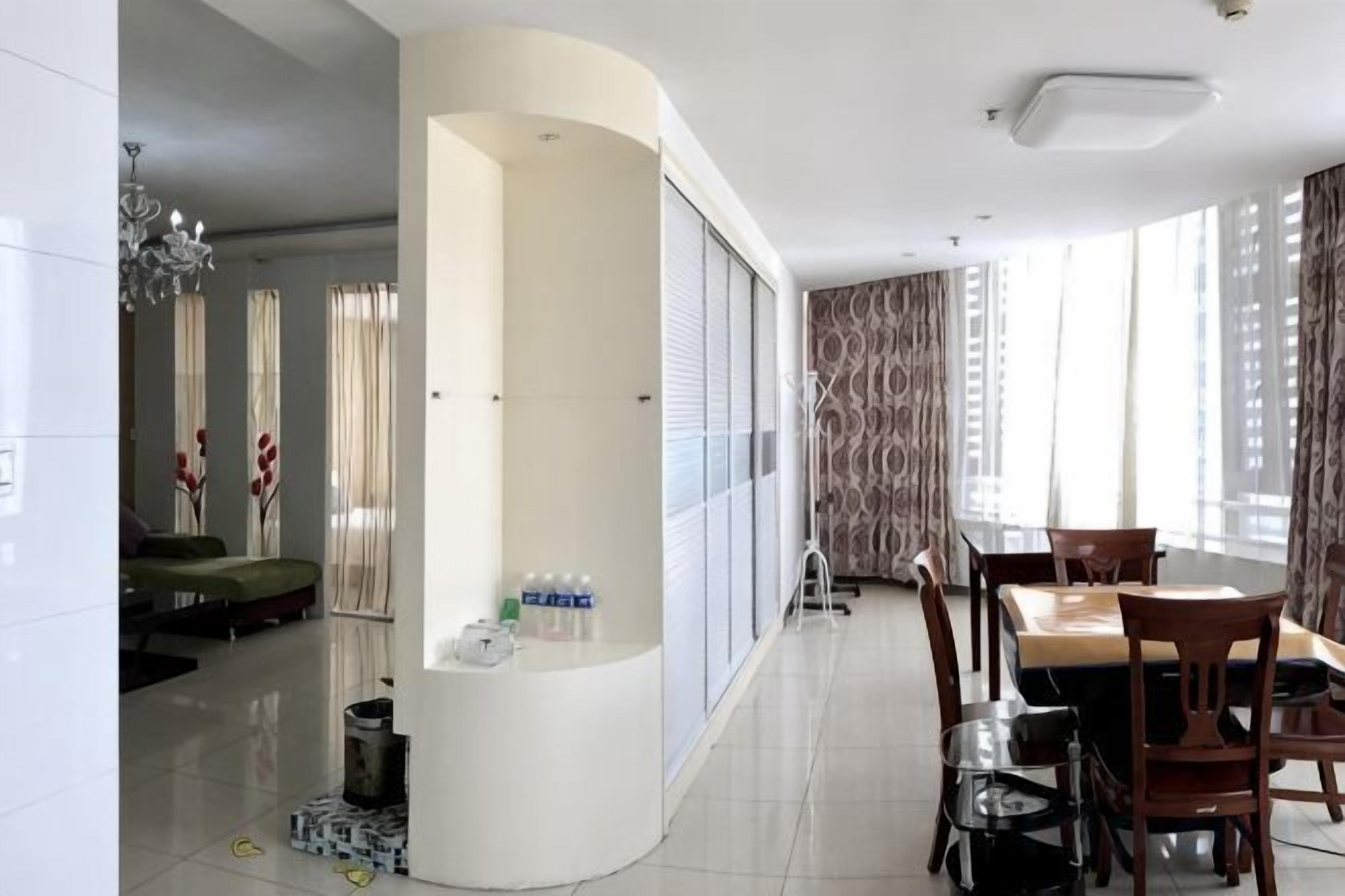 Habitación de hotel con un separador blanco curvo, una mesa de comedor con sillas, un sofá y una pequeña zona de cocina.