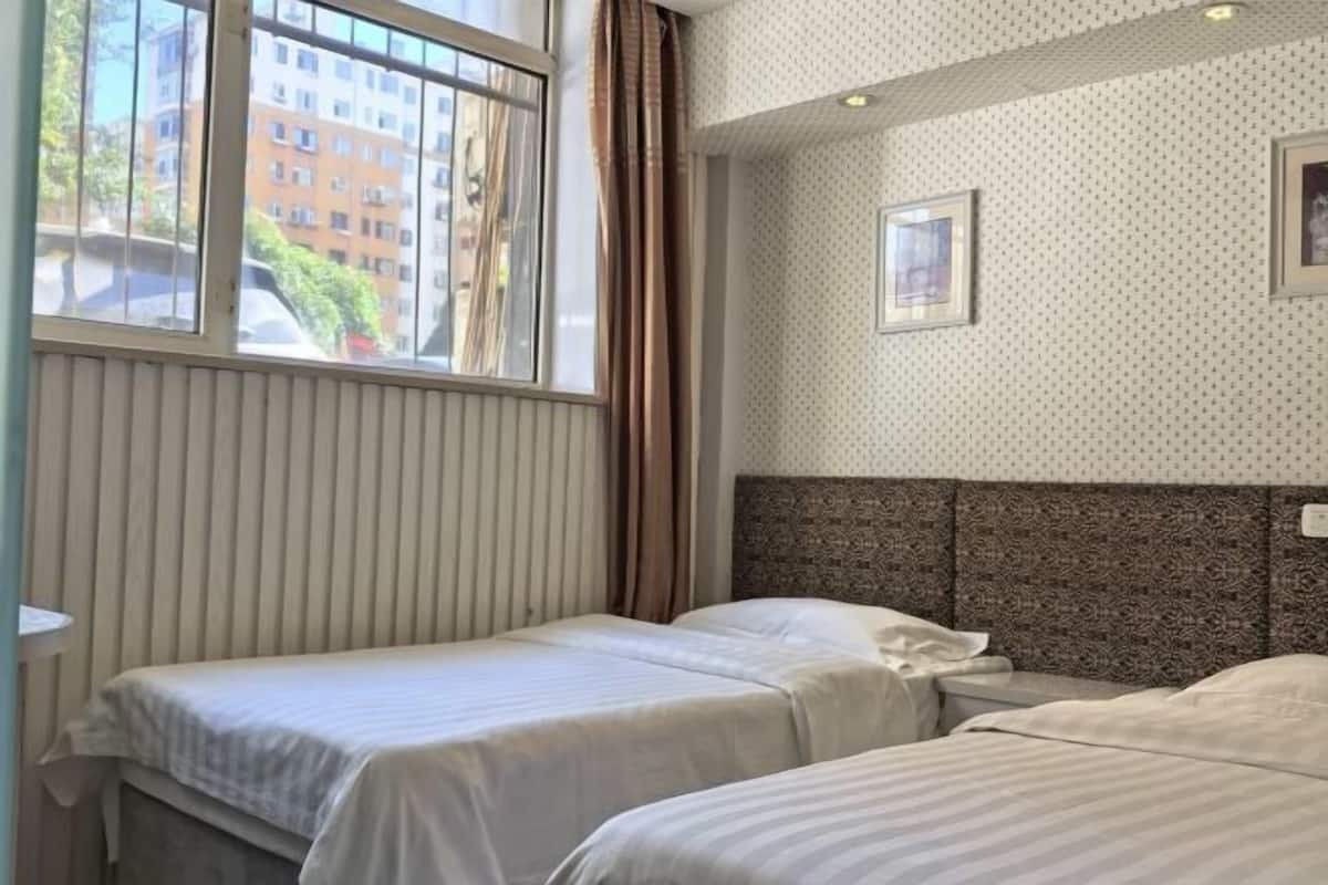 Une chambre d’hôtel avec deux lits, une fenêtre avec des rideaux, un papier peint à motifs et un tableau encadré au mur.