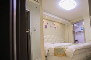 A bedroom with a white tufted headboard, a bed with white bedding and a decorative throw, a wall-mounted light fixture, and a door leading to a bathroom. - Shell Chain Hotel (Mudanjiang North Railway Station Guangsha Branch) (Mudanjiang)