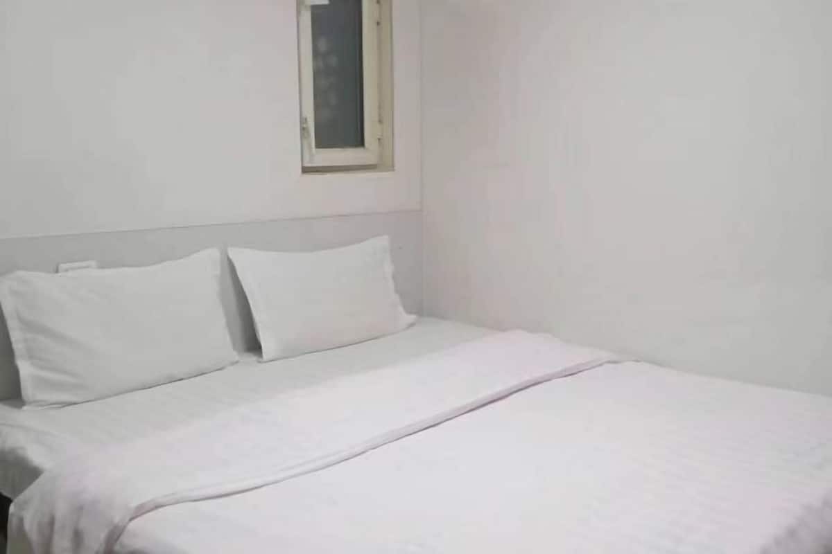 Une chambre d’hôtel simple et sans fioritures, avec un lit simple, du linge de lit blanc et une petite fenêtre.