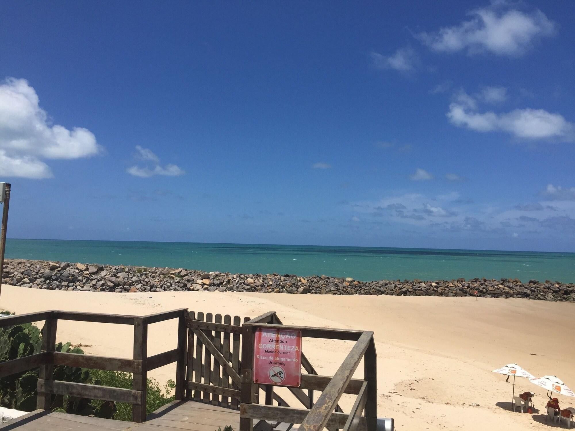 A beach with a wooden walkway leading to the shore, a sign warning of marine currents and drowning risk, and a clear view of the ocean.