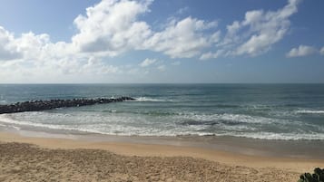 Uma praia com um paredão rochoso, areal arenoso e céu claro com nuvens.