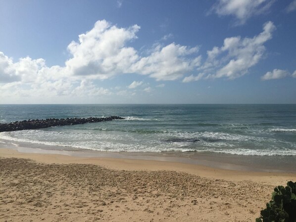 A beach with a rocky breakwater, sandy shore, and a clear sky with clouds.