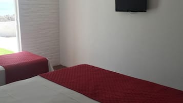 Quarto com duas camas, uma com colcha vermelha e outra com colcha branca. Há um objeto preto na parede, possivelmente um alto-falante ou uma tela pequena.