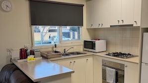 A compact kitchen with white cabinets, a microwave, a stove, and a sink. There's a clock on the wall and a window above the sink.
