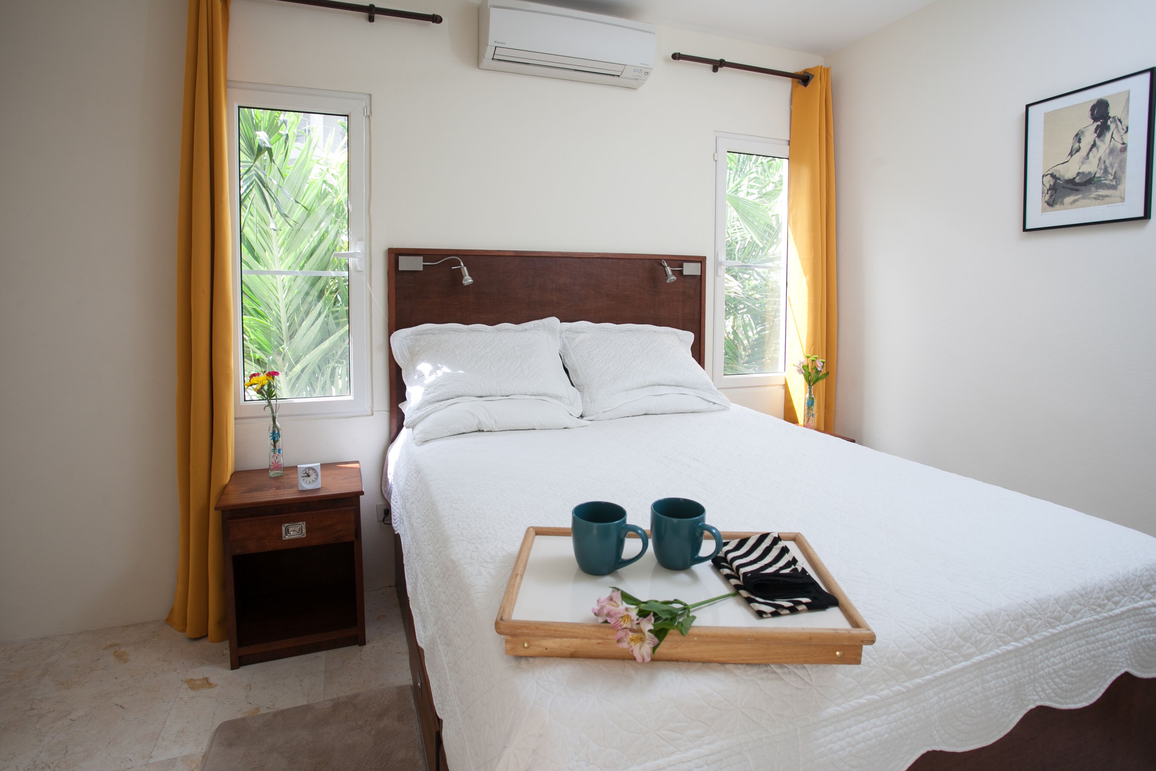 A neatly made bed with a tray of items, a bedside table with a vase, and a window with curtains.