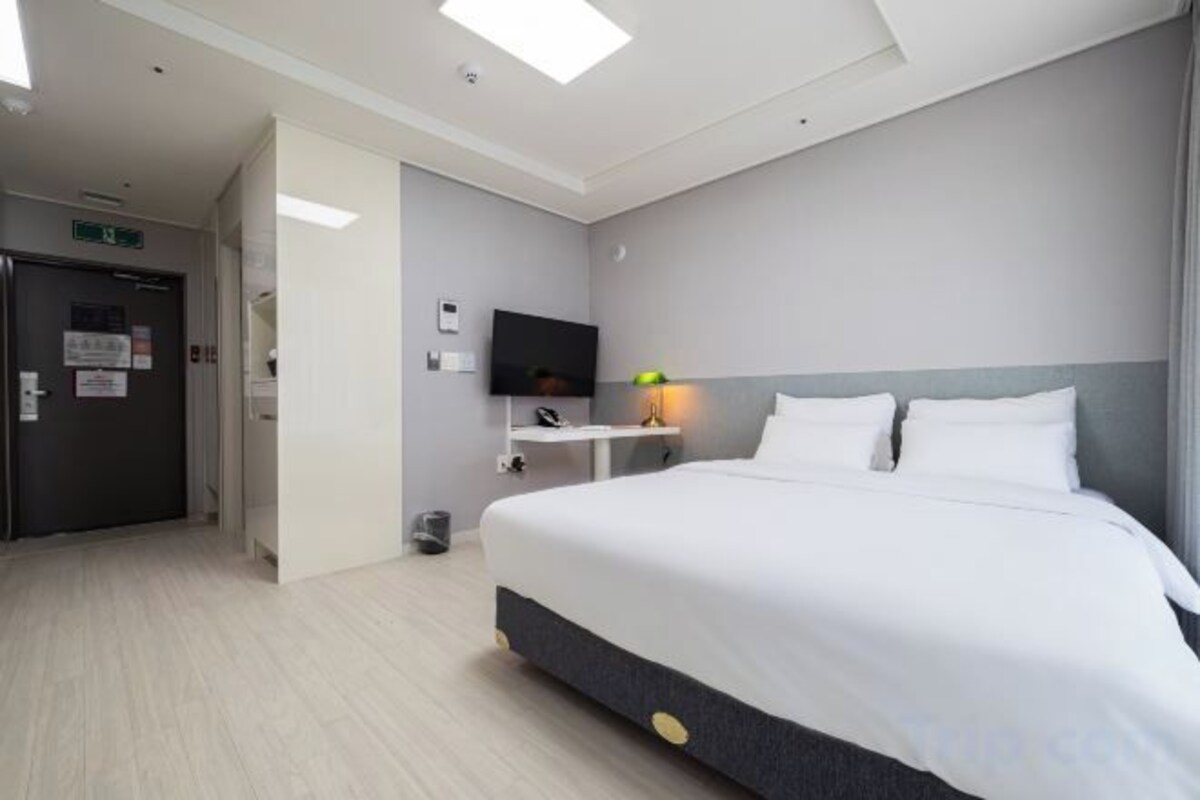 A hotel room with a bed, a television, a small table with a lamp, and a door.