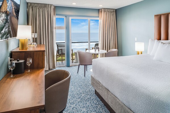A hotel room with a bed, a desk with a lamp, a chair, a table with a view of the ocean, and a balcony with curtains.