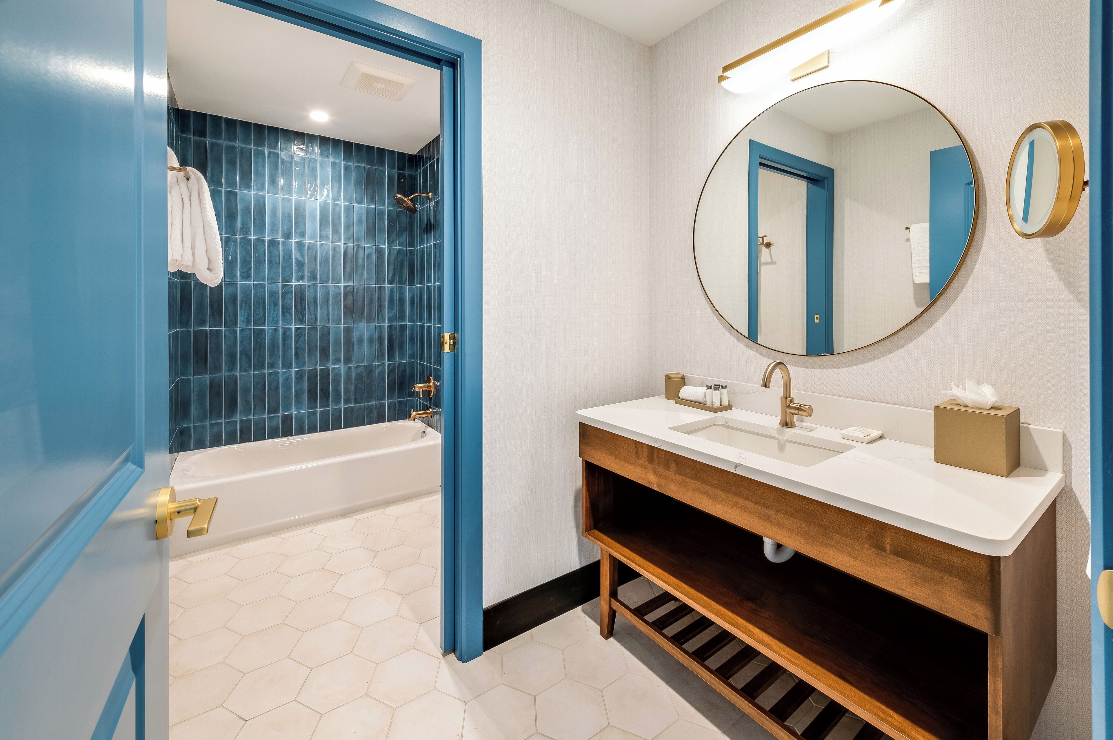 A bathroom with a blue-tiled shower, a white sink with a wooden vanity, and a round mirror with a gold frame.