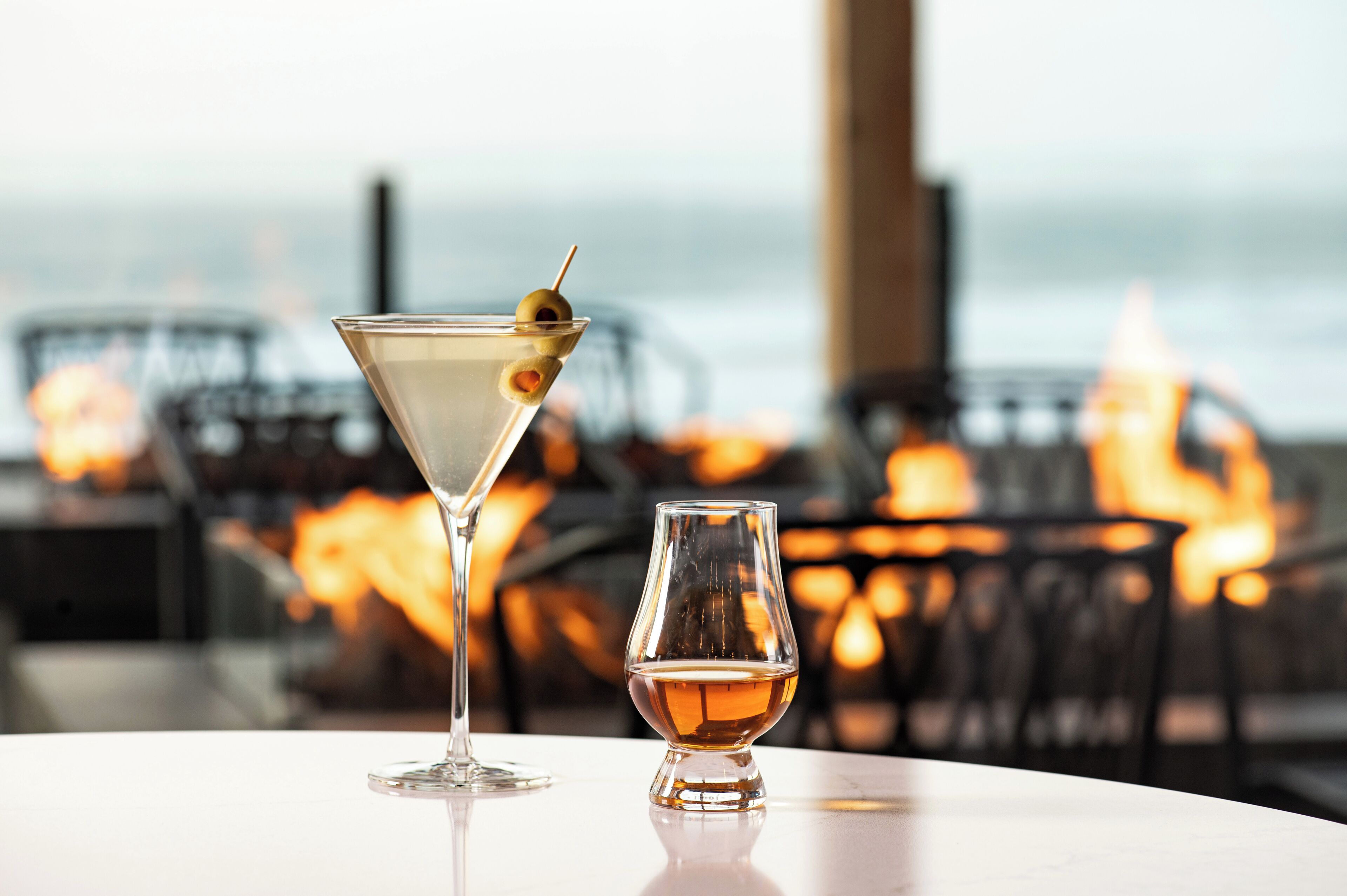 A martini glass and a small glass with a drink on a table with a view of a fire and a building in the background.