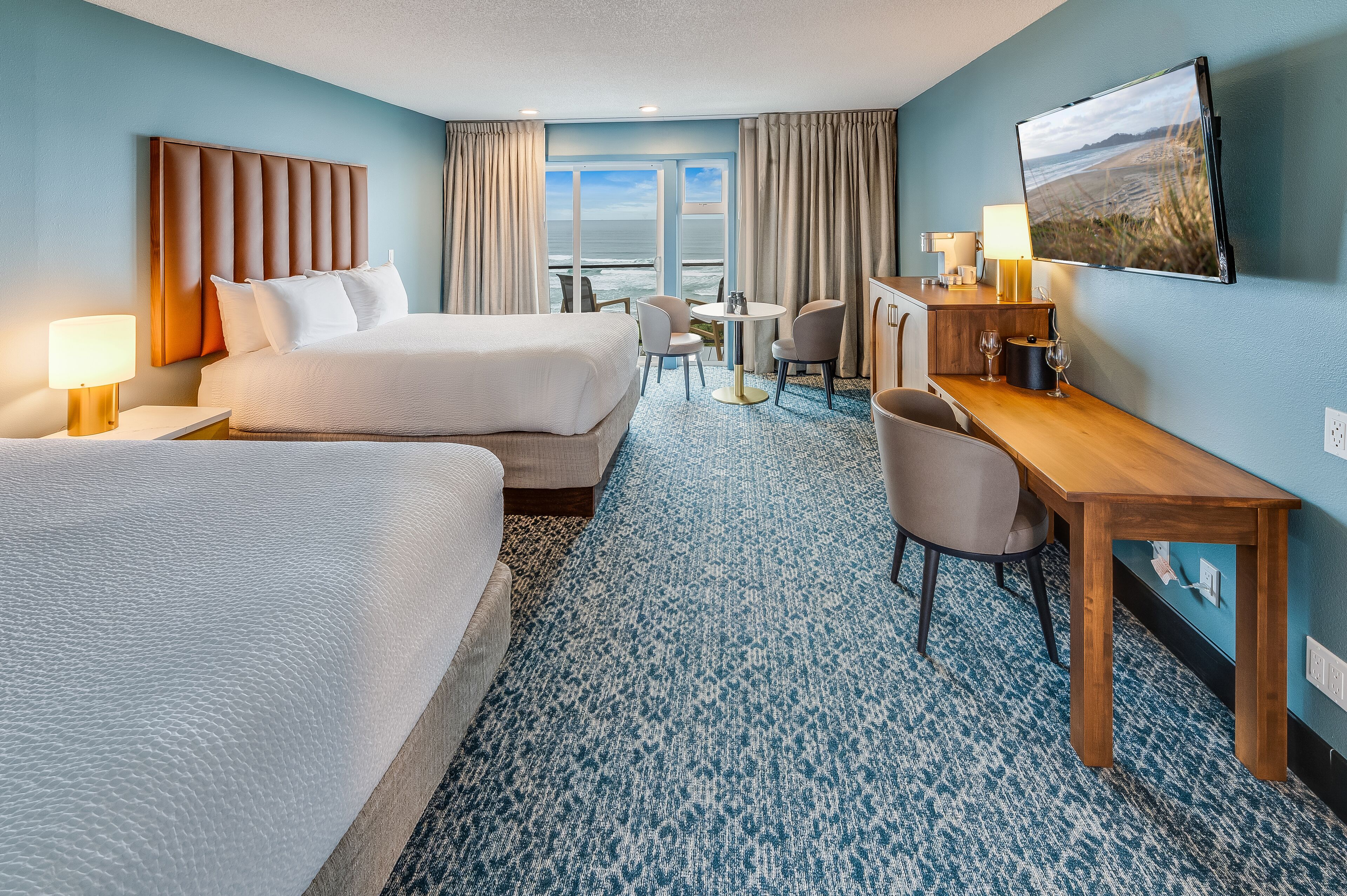 A hotel room with two beds, a desk, a chair, a table, and a large window offering a view of the beach.