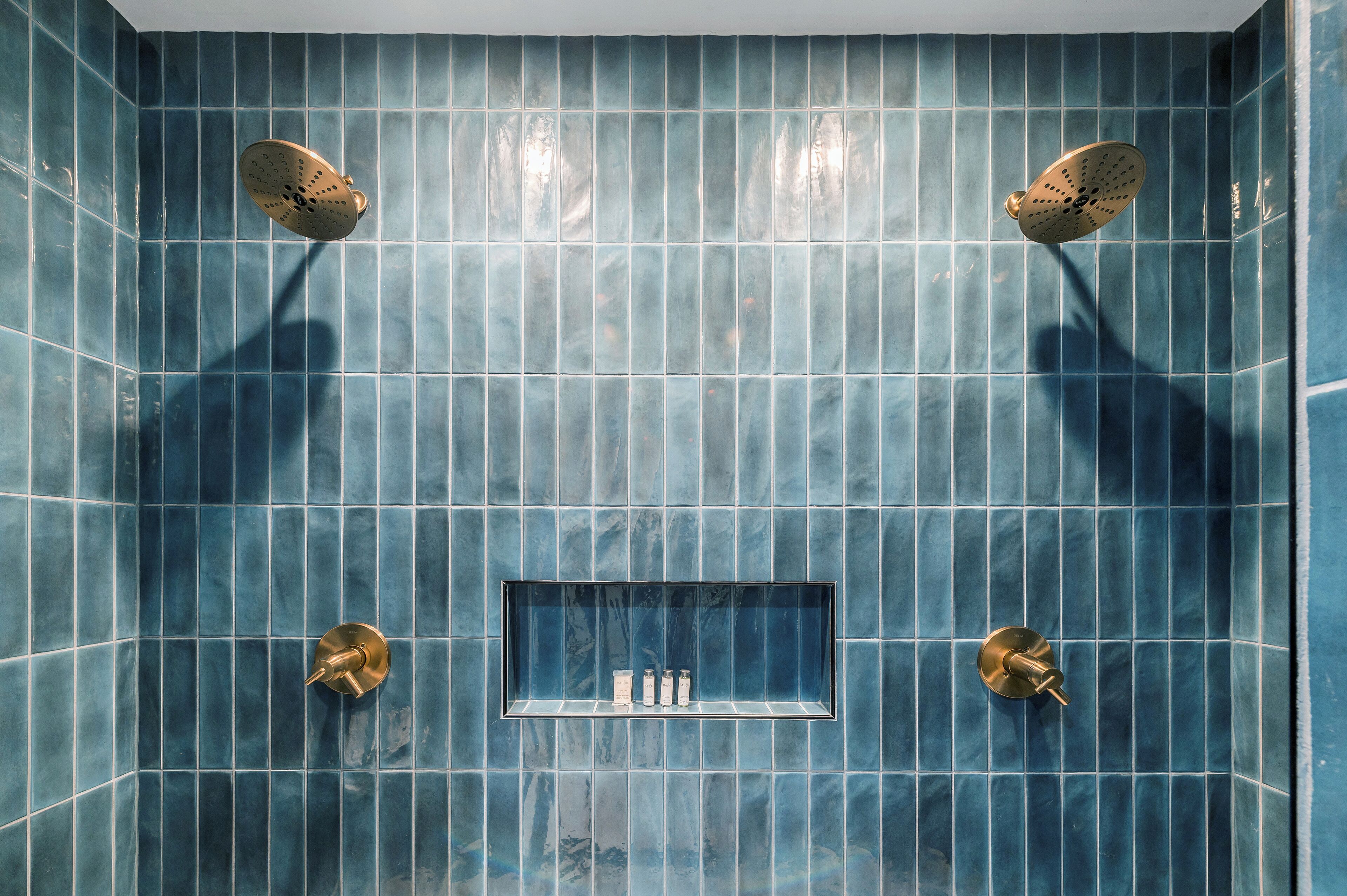 A bathroom with blue tiled walls, a rectangular shelf with toiletries, and two showerheads.
