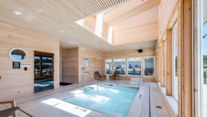 A modern indoor swimming pool with wooden walls and ceiling, clear glass doors, and a view of the outside.