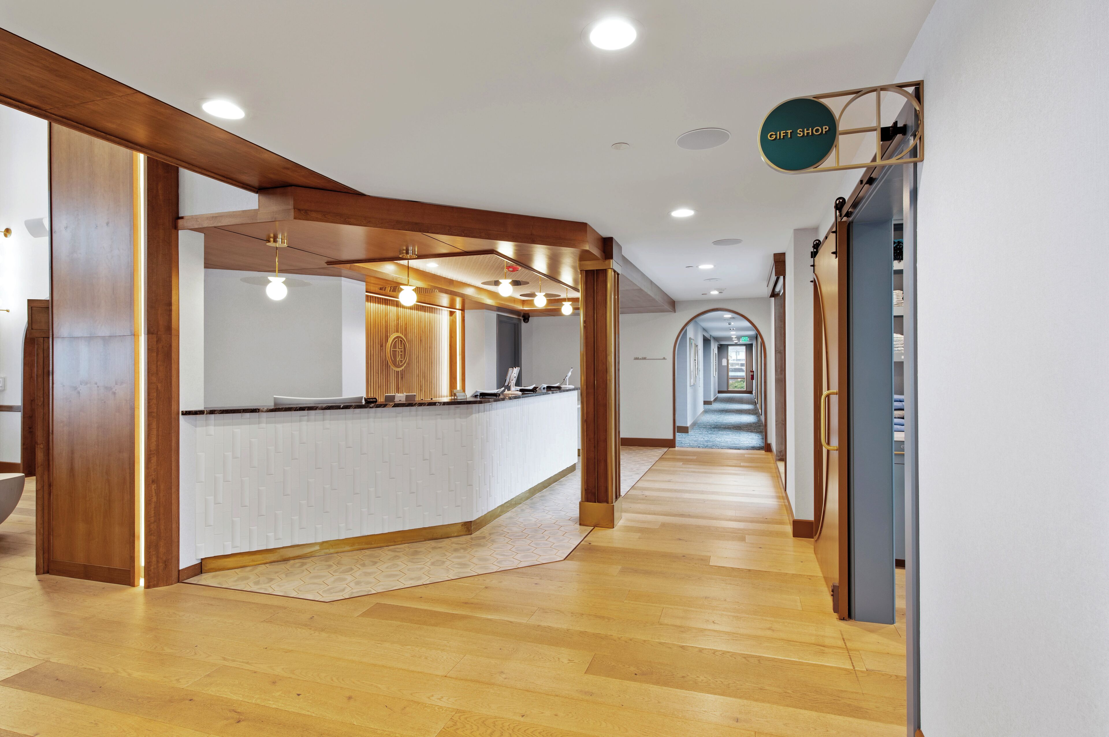 A modern hotel lobby with a wooden reception desk, a gift shop sign, and a hallway leading to other areas.