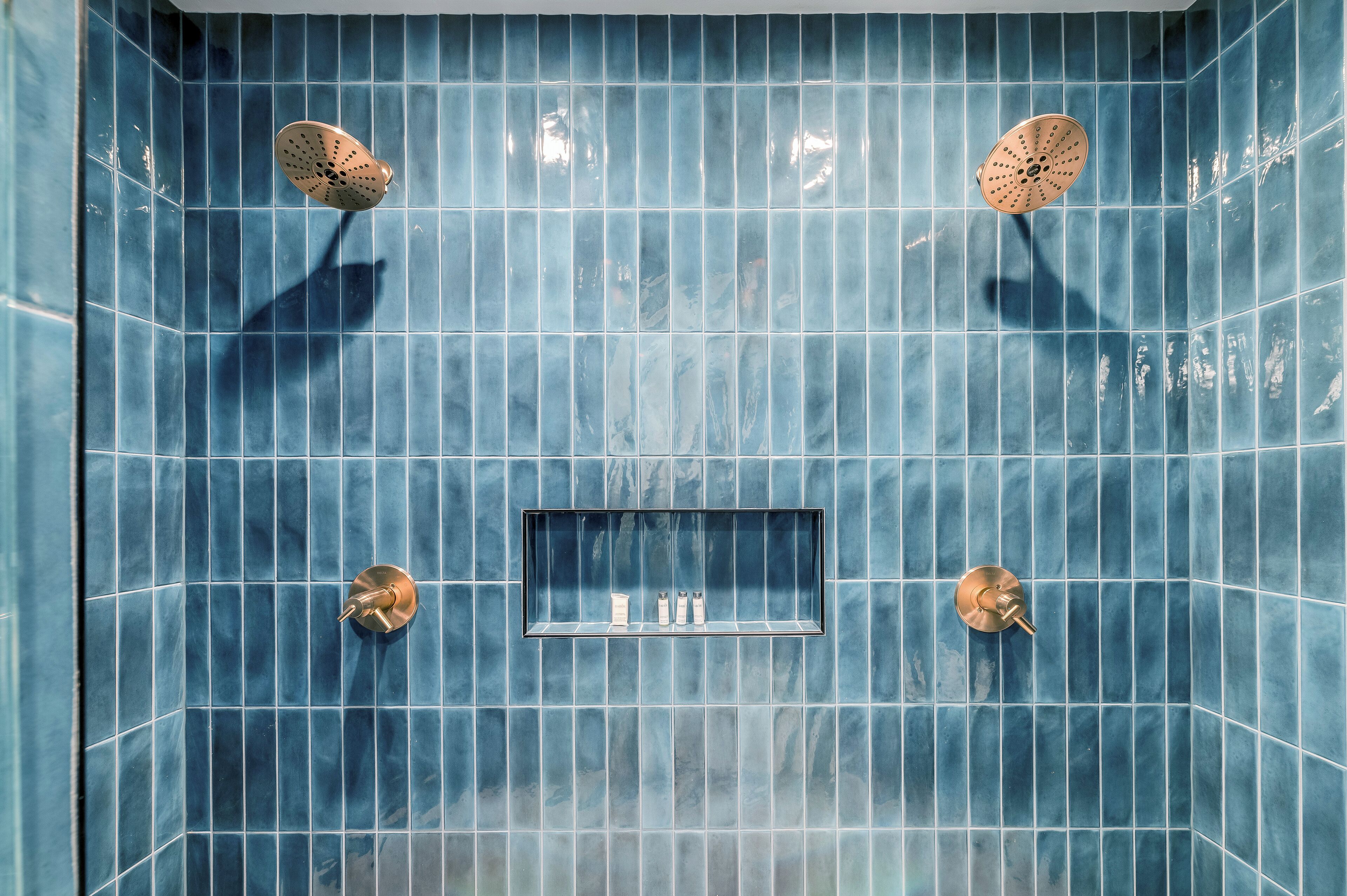 A bathroom with blue tiles, two showerheads, and a shelf with toiletries.